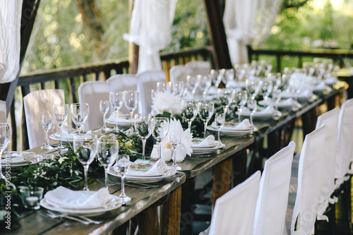 Decorated elegant wooden table for a wedding feast in the gazebo in the rustic style with eucalyptus and flowers, porcelain plates, glasses, white chairs