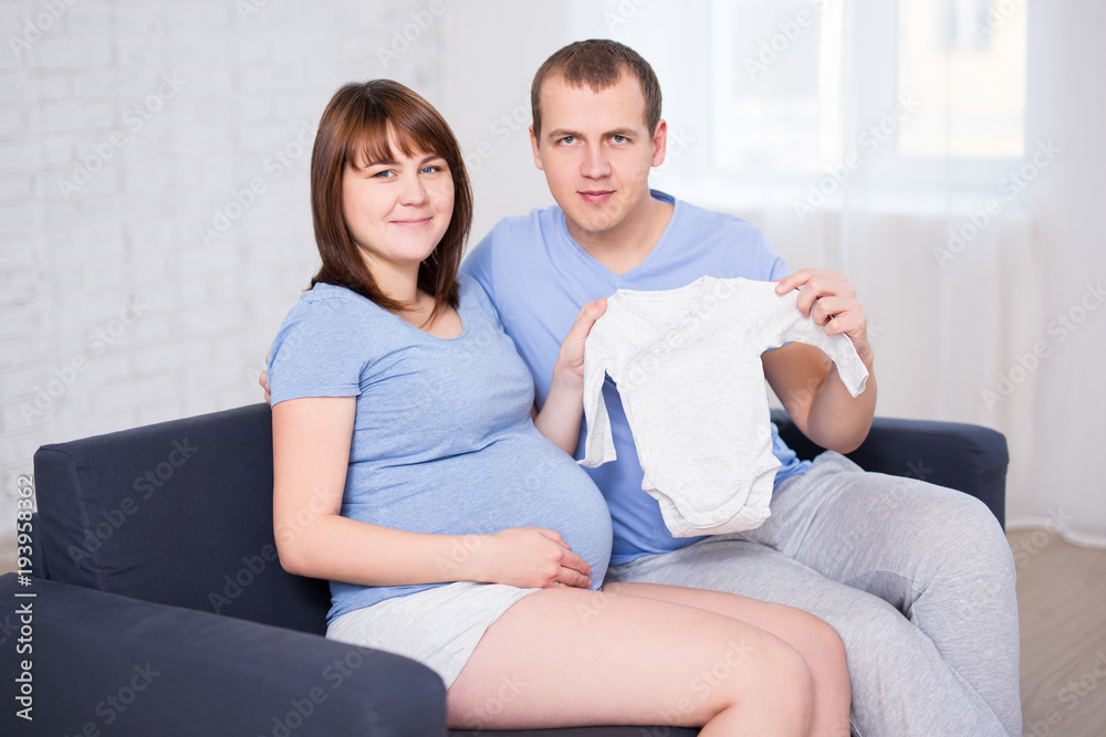 young beautiful pregnant couple sitting on sofa in living room and holding baby bodysuit young beautiful pregnant couple sitting on sofa in living room and holding baby bodysuit