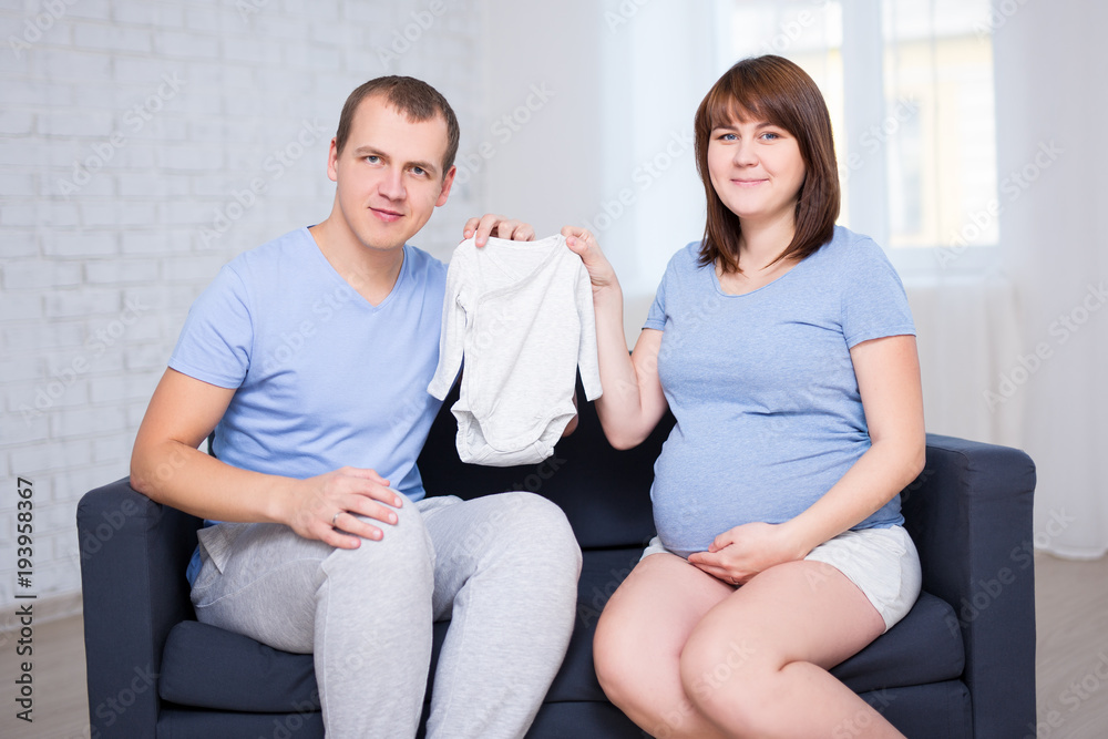 family concept - beautiful pregnant couple sitting on sofa in living room and holding baby bodysuit family concept - beautiful pregnant couple sitting on sofa in living room and holding baby bodysuit