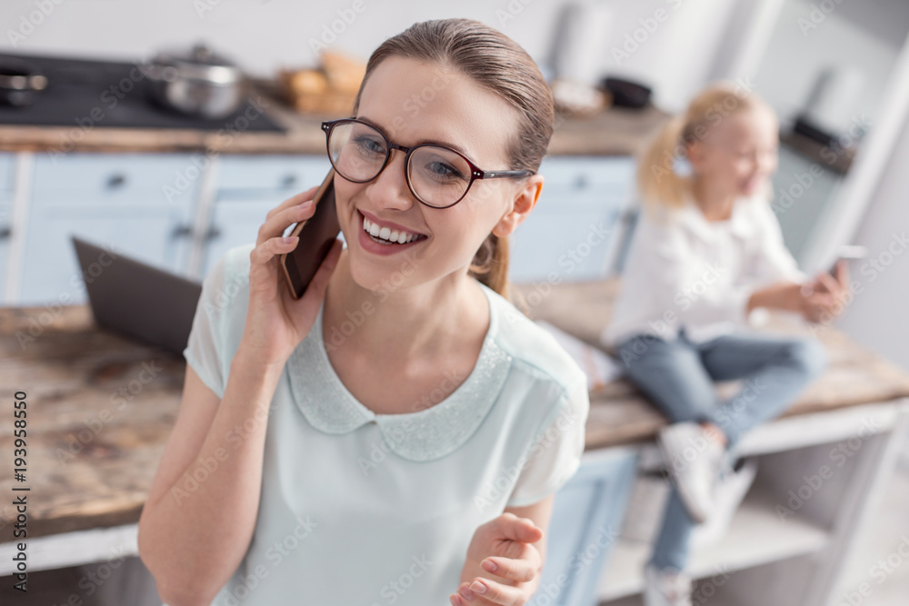 Fototapeta premium Great news. Appealing vigorous enthusiastic mom chatting on phone while laughing and wearing glasses