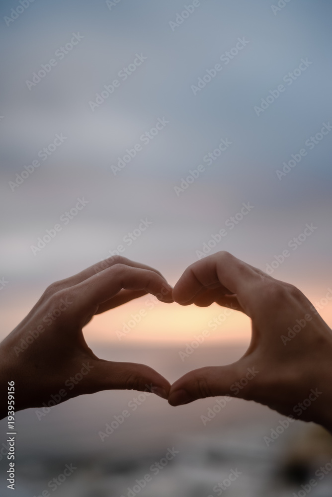 Fototapeta premium Bright sunset and hands of a man forming a heart shape silhouette on natural oudoors abstract background. Close up on emotional human feeling, over sunny seascape shore leisure travel landscape