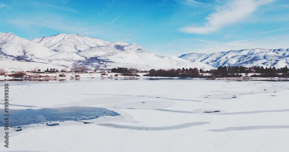 Aerial view flying over winter lake covered in ice with snow covered mountains in background Aerial view flying over winter lake covered in ice with snow covered mountains in background