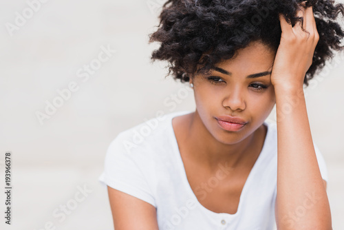 Beautiful young black woman with sad, pensive, reflective look, against white wall background with copy space for your text or advertising content. Portait of worry dark skinned female in white tshirt