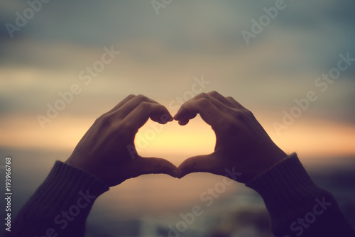 Bright sunset and hands of a man forming a heart shape silhouette on natural oudoors abstract background. Close up on emotional human feeling, over sunny seascape shore leisure travel landscape