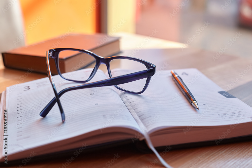 Glasses and a pen on a diary on a wooden table next to a book Glasses and a pen on a diary on a wooden table next to a book