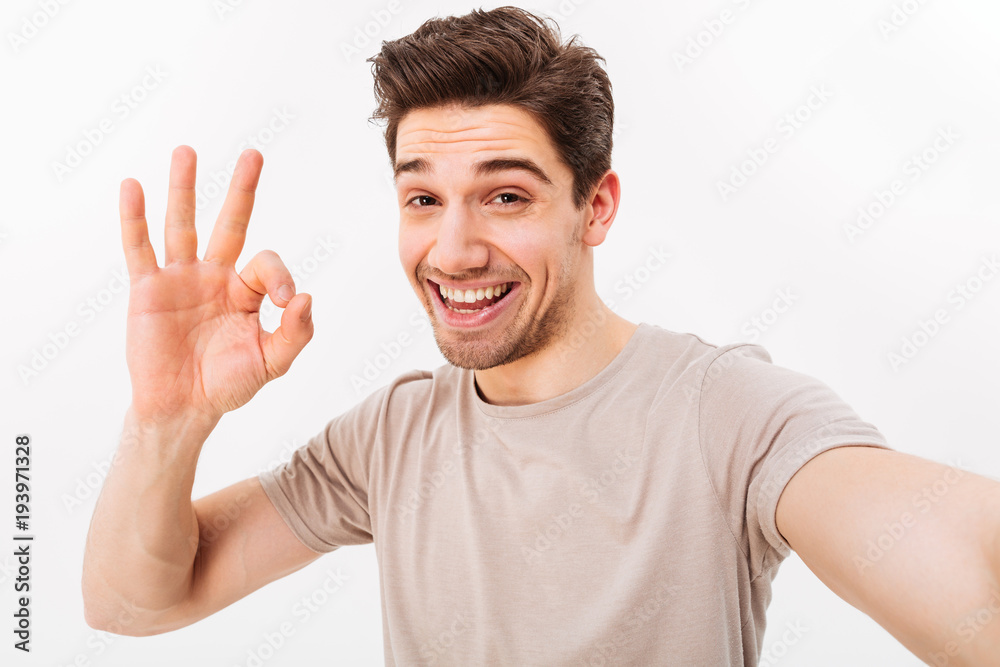 Cheerful man in casual t-shirt and bristle on face smiling on camera with alright sign while taking selfie, isolated over white background Cheerful man in casual t-shirt and bristle on face smiling on camera with alright sign while taking selfie, isolated over white background