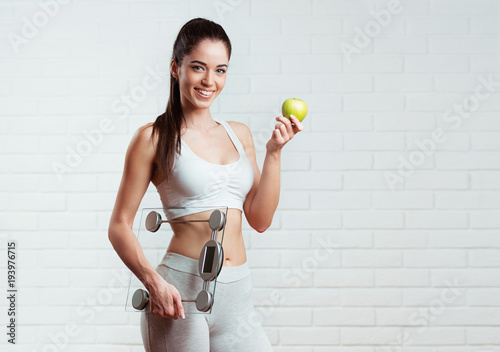 Beautiful, fit, young woman holding a weight scale, over a white brick wall