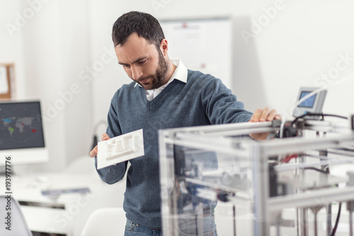 Nice result. Handsome dark-haired young man holding a recently printed 3D model and examining it thoroughly while resting his hand on a 3D printer