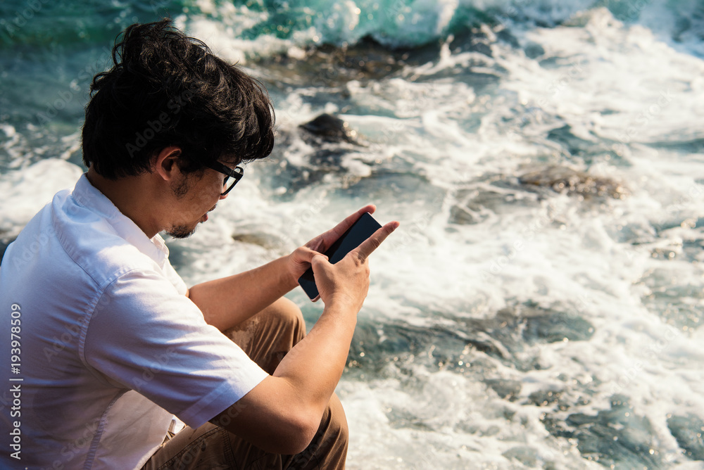 Asian man using phone typing message to chatting in social network while sitting on rock on sea.
