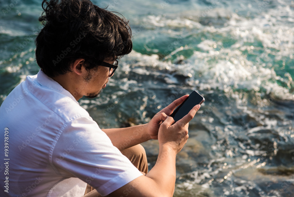 Asian man using phone typing message to chatting in social network while sitting on rock on sea.