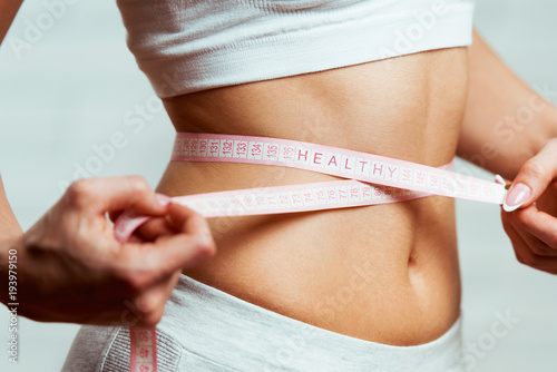 Beautiful, fit, young woman measuring her waist with a measuring tape, with word HEALHTY written on it, close up