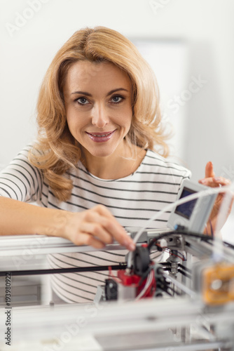 High technology. Pleasant young woman posing for the camera and smiling while touching the part of a 3D printer before printing something on it