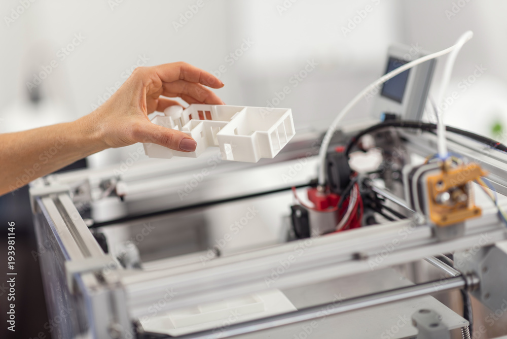 Best replica. The close up of a delicate female hand removing a house model from a 3D printer, having printed it for her project