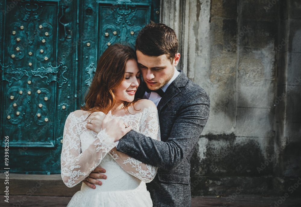 Wedding couple hugs near the vintage green door. Stone walls in ancient town background. Rustic bride with hair down in lace dress and groom in grey suit and bow tie. Tender embrace. Romantic love.