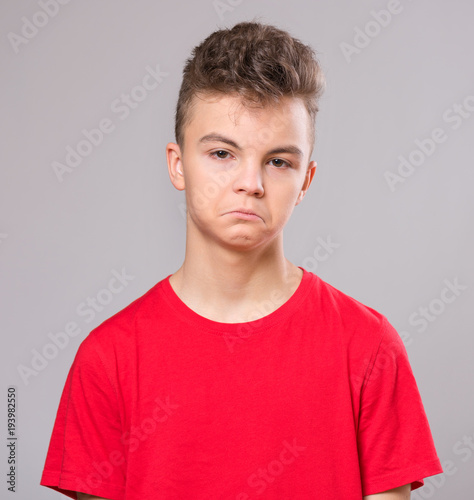 Emotional portrait of caucasian upset problem teen boy. Sad boy looking at camera. Worried child wearing red t-shirt, on gray background.