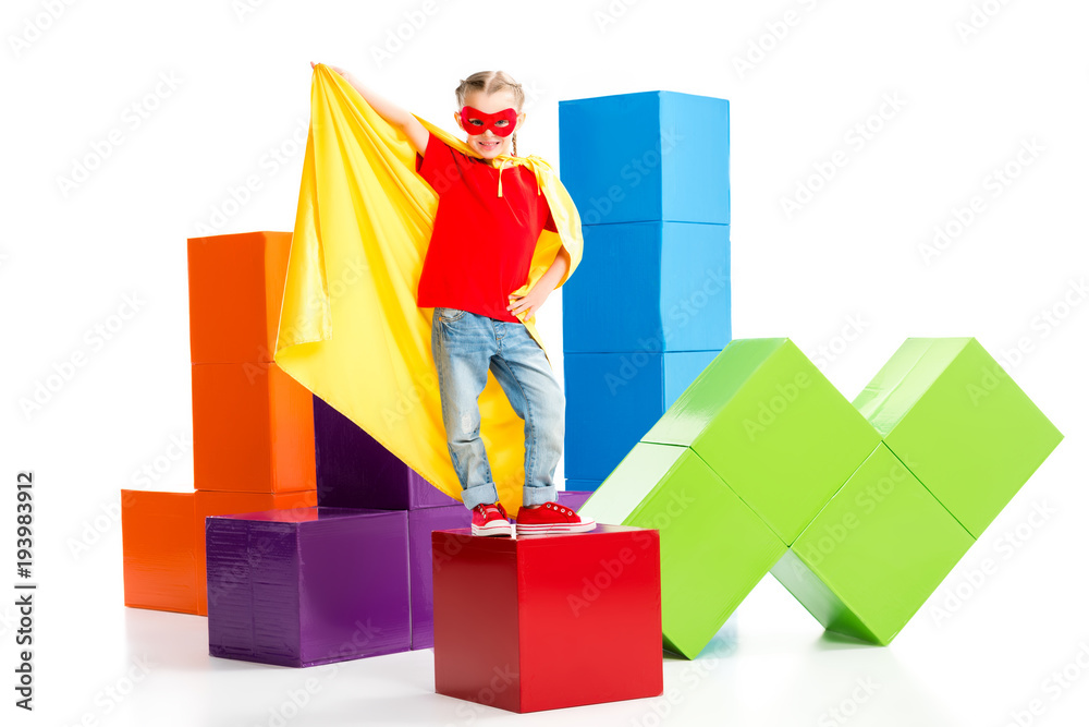 Smiling supergirl wearing yellow cape and red mask for eyes standing on cube isolated on white Smiling supergirl wearing yellow cape and red mask for eyes standing on cube isolated on white