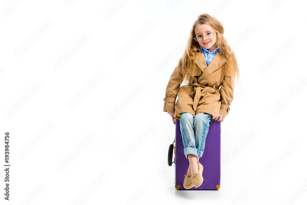 Stylish child in glasses and trench coat sitting on suitcase isolated on white Stylish child in glasses and trench coat sitting on suitcase isolated on white