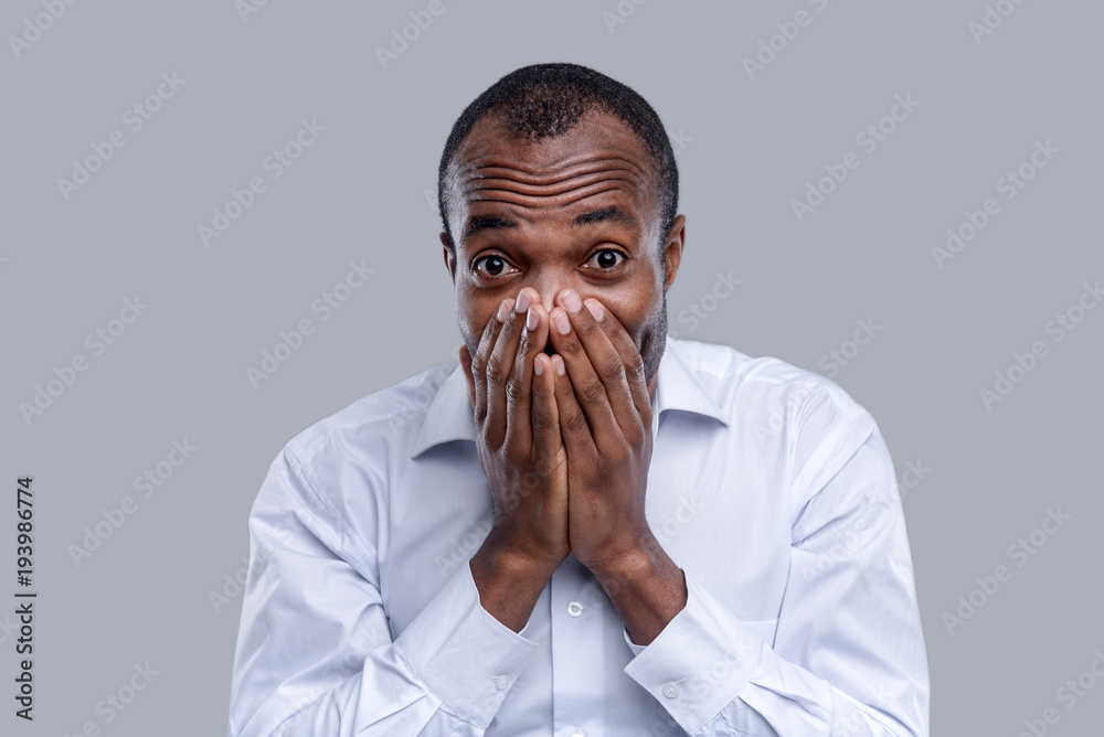 Surprised. Good-looking amazed dark-eyed afro-american man having round eyes and wearing a white shirt and closing his mouth with his hands Surprised. Good-looking amazed dark-eyed afro-american man having round eyes and wearing a white shirt and closing his mouth with his hands