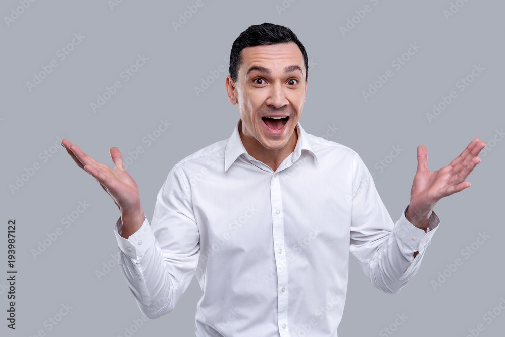 Inspiration. Handsome exuberant dark-eyed young man wearing a white shirt and smiling and having his mouth opened and arms up Inspiration. Handsome exuberant dark-eyed young man wearing a white shirt and smiling and having his mouth opened and arms up