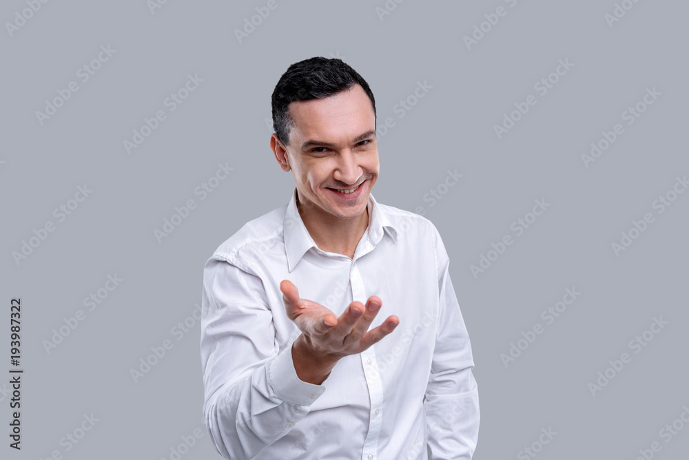 Making an offer. Handsome cheerful dark-eyed young man wearing a white shirt and smiling and outstretching his arm Making an offer. Handsome cheerful dark-eyed young man wearing a white shirt and smiling and outstretching his arm