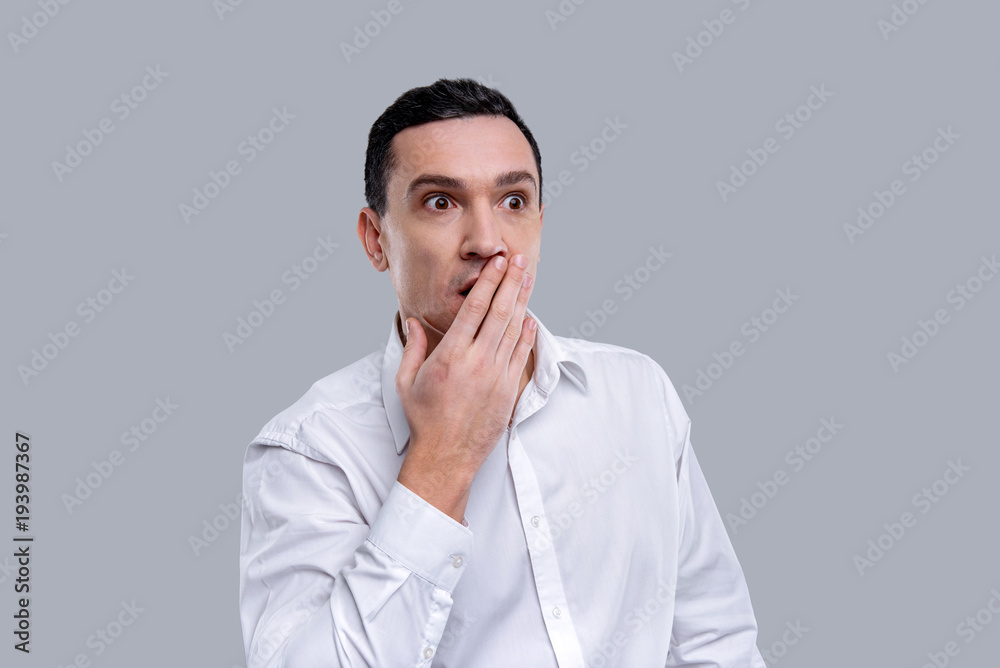 Baffled. Attractive amazed dark-eyed young man wearing a white shirt and having his eyes wide-opened and closing his mouth with a hand Baffled. Attractive amazed dark-eyed young man wearing a white shirt and having his eyes wide-opened and closing his mouth with a hand