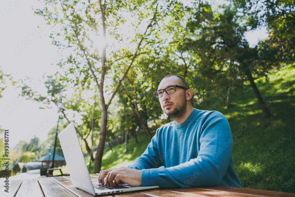 Young successful smart man businessman or student in casual blue shirt glasses sitting at table with mobile phone in city park using laptop working outdoors on green nature. Mobile Office concept. Young successful smart man businessman or student in casual blue shirt glasses sitting at table with mobile phone in city park using laptop working outdoors on green nature. Mobile Office concept.