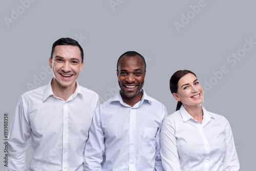 Amiable atmosphere. Happy young dark haired co-workers smiling and wearing white shirts and standing according to height