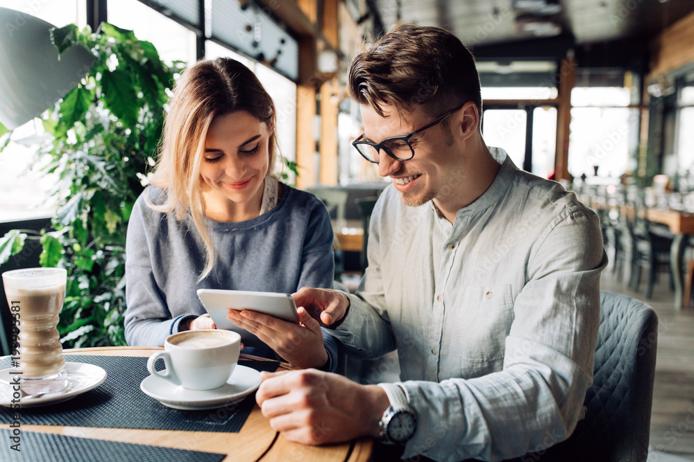 Cheerful man and beautiful woman resting together at cafe while looking at tablet screen, drinking coffee.