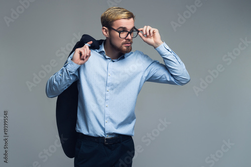Confident and brutal. Business man holding the jacket on the shoulder by one hand and putting modern glasses on eyes by the other in front of grey background.