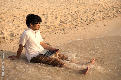 Asian man with his computer on the beach with waves from sea and golden sunlight at evening. working on computer at outdoors while traveling at sea in summer holiday. freelancer work concept.