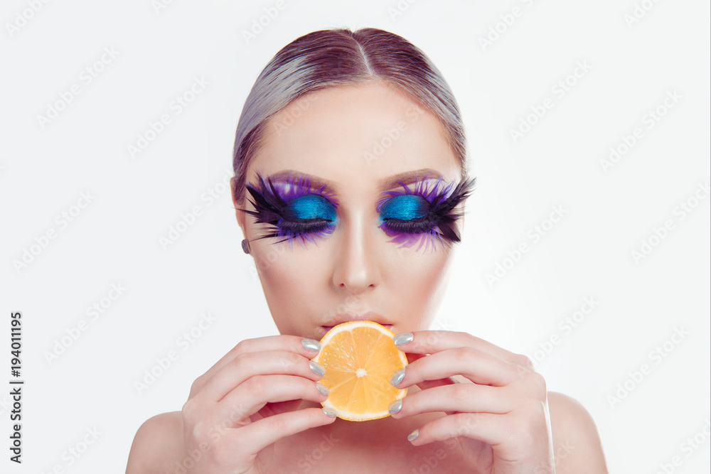 Woman with artistic purple blue eyes makeup feather on eyelashes holding lemon slice in mouth Woman with artistic purple blue eyes makeup feather on eyelashes holding lemon slice in mouth