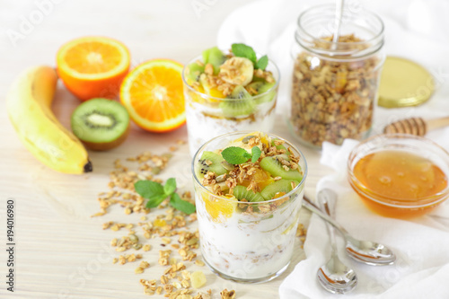 Tasty and healthy breakfast or snack two glasses of yogurt, granola and kiwi, orange, banana slices, honey, mint leaves on a white wooden background.
