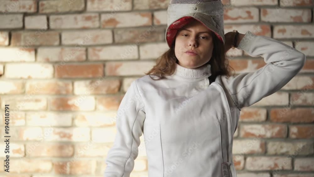 Fencer girl is pulling mask with metal protection grid on a head. She is standing in a gym for training, she is looking on a camera, medium shot Fencer girl is pulling mask with metal protection grid on a head. She is standing in a gym for training, she is looking on a camera, medium shot