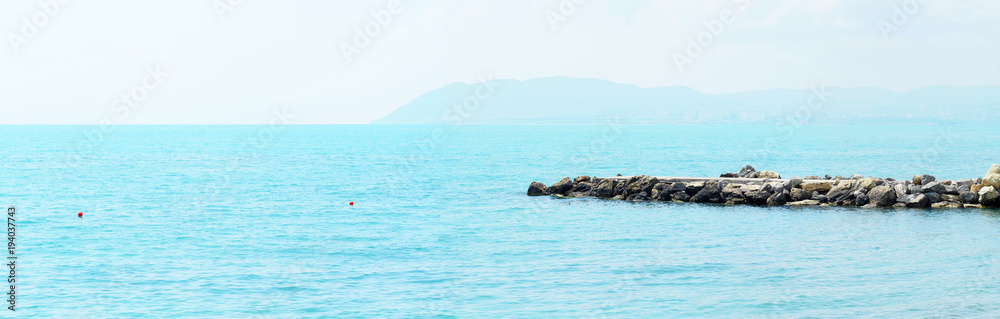 Fototapeta premium Sea panoramic landscape, stone breakwater. Sunny morning summer landscape. Rocky coast and a seawall. Breakwater in blue sea. Skyline, sea line