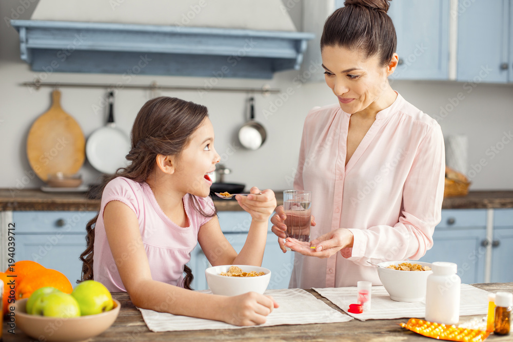 Great day. Beautiful exuberant dark-haired young mother smiling and giving vitamins to her daughter and the girl having breakfast and sitting at the table Great day. Beautiful exuberant dark-haired young mother smiling and giving vitamins to her daughter and the girl having breakfast and sitting at the table