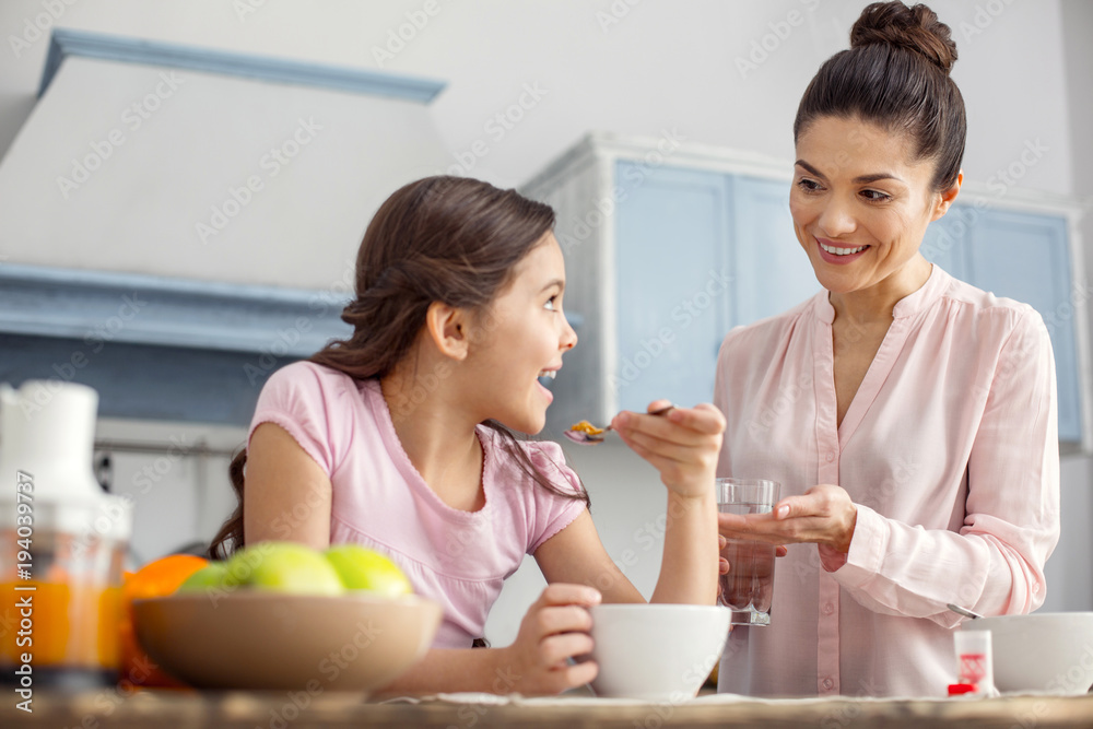 Good mood. Pretty happy dark-haired young mother smiling and giving vitamins to her daughter and the girl having breakfast and sitting at t Good mood. Pretty happy dark-haired young mother smiling and giving vitamins to her daughter and the girl having breakfast and sitting at t