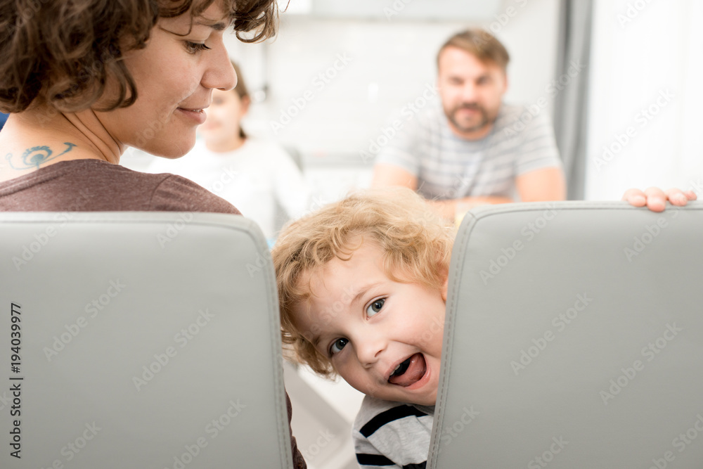 Portrait of playful little boy peeking out sitting at table in dining room and making faces while waiting for dinner Portrait of playful little boy peeking out sitting at table in dining room and making faces while waiting for dinner
