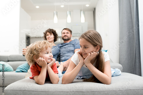 Portrait of happy family with two kids enjoying evening watching TV together sitting on sofa in living room, focus on brother and sister in foreground