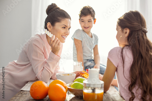 Good vitamins. Attractive smiling dark-haired young mother holding vitamins and talking with her kids about healthcare and the boy sitting on the table