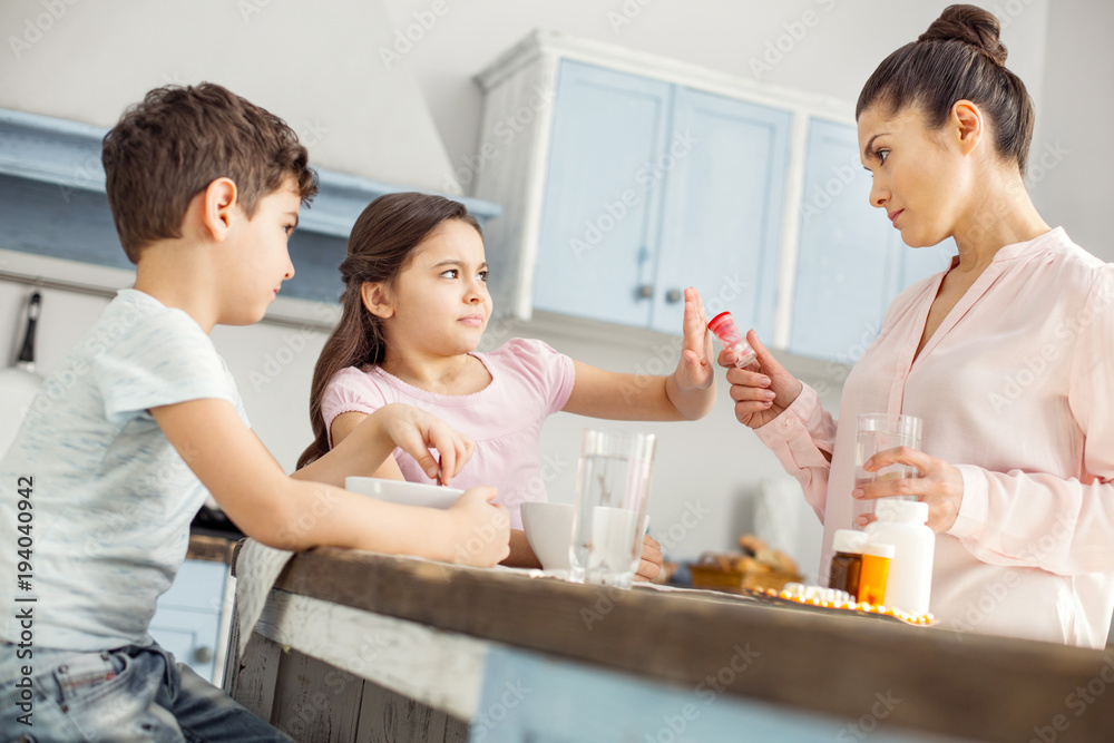 I hate them. Lovely annoyed dark-haired little girl having breakfast with her brother and refusing to take vitamins and her mom looking strictly I hate them. Lovely annoyed dark-haired little girl having breakfast with her brother and refusing to take vitamins and her mom looking strictly