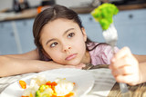 I feel sad. Pretty sad dark-haired little girl having healthy breakfast and looking at the green vegetable on her fork and not liking it