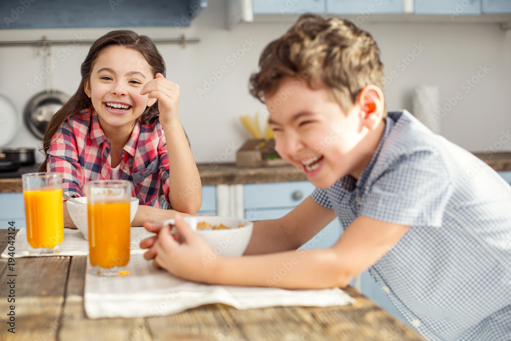 Having fun together. Pretty joyful little dark-haired girl laughing and having breakfast with her brother and her brother smiling too Having fun together. Pretty joyful little dark-haired girl laughing and having breakfast with her brother and her brother smiling too