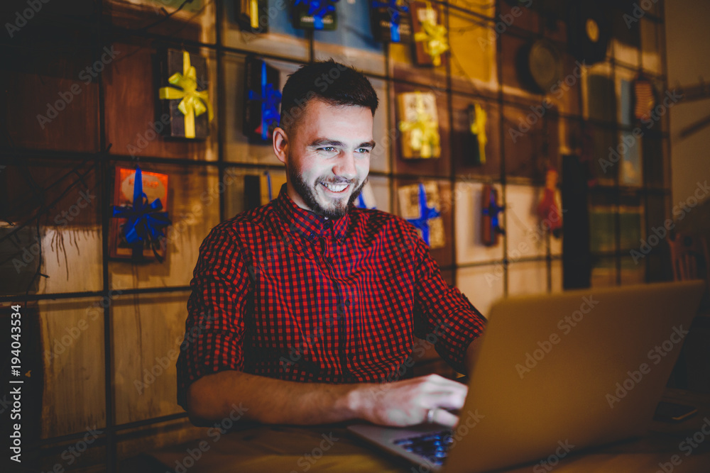 Theme training and computers. A young man with a beard and in a shirt uses a laptop, prints on the keyboard in a coffee shop at a wooden table in the evening. Christmas decor and hang a garland Theme training and computers. A young man with a beard and in a shirt uses a laptop, prints on the keyboard in a coffee shop at a wooden table in the evening. Christmas decor and hang a garland