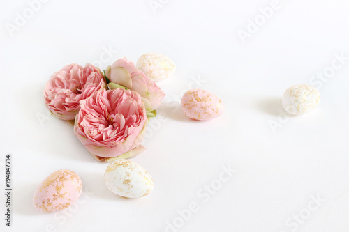Spring greeting card, invitation. Pink and white Easter eggs with golden spots and rose flowers lying on white table. Feminine styled stock photo, floral composition.