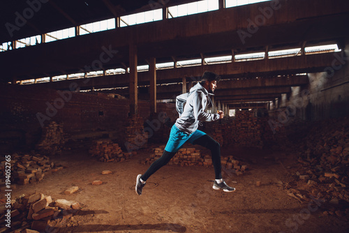 Portrait of active motivated afro-american young attractive athletic man with earphones running inside of the abandoned place.