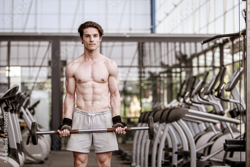 A shirtless young muscular man working out in gym doing exercises with equipment in fitness center.