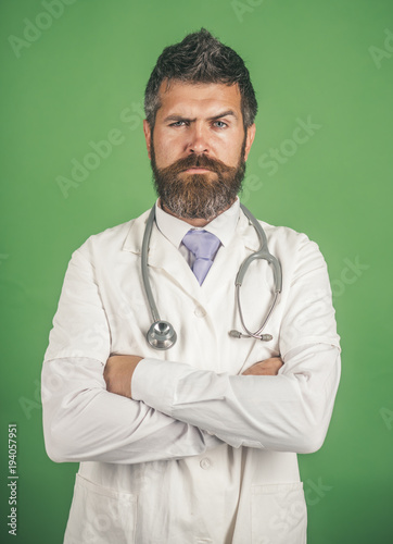 Healthcare, medicine, pharmacy and treatment concept - serious male doctor in white medical gown. Handsome bearded physician with stethoscope. Isolated on green background.