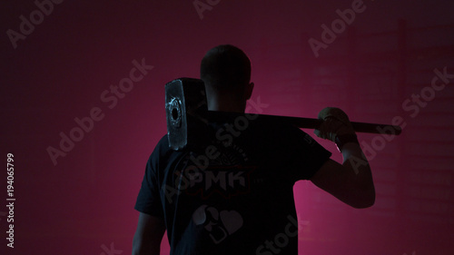 Man with a sledgehammer behind his back. Guy with a sledgehammer in the red room. Man with a big hammer or a sledgehammer behind comes into the room with a red light. Muscular man with hammer from