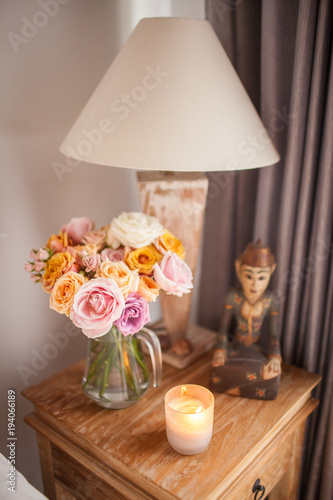 Wooden Bed with White Sheets. A Bedside Table by the Bed with a Lamp, a Bouquet of Flowers from Roses, Candle and Hand Cream