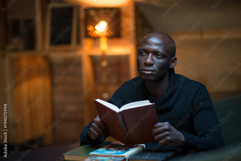 A young Arican man is reading a book in a room at a table. student, businessman
